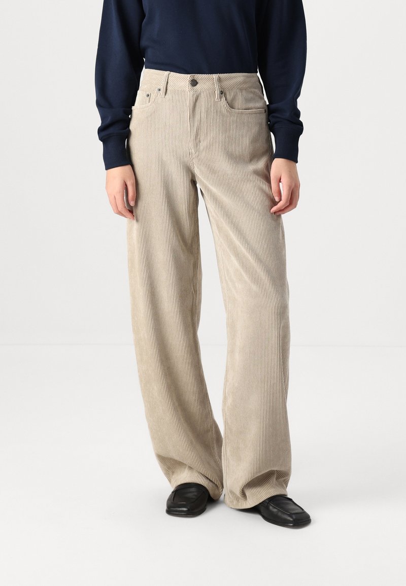 Personne portant un pantalon large en velours côtelé beige taille haute, des mocassins noirs et un haut à manches longues foncé, debout sur un fond blanc.