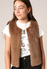 Gilet en fausse fourrure marron sans manches, doté d'une texture douce et d'une ouverture sur le devant, porté sur un t-shirt graphique blanc et un pantalon noir.