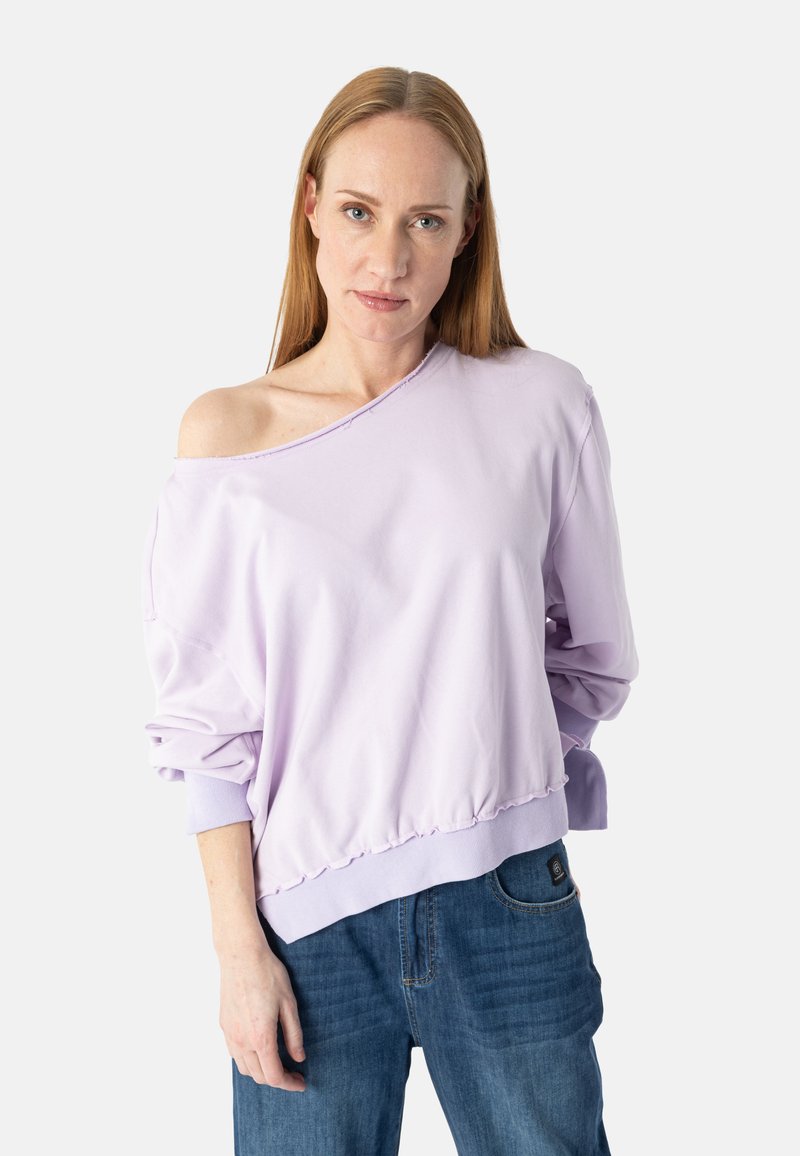 Femme aux cheveux rouges lisses portant un sweat-shirt lavande ample à épaules dénudées et un jean bleu, debout devant un fond clair uni.
