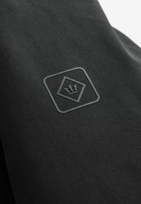 Juodas audinys su lygiu paviršiumi turi kvadratinį iškilų logotipą su karūna viduje, kuris išdėstytas akcentuotoje vietoje paviršiuje.