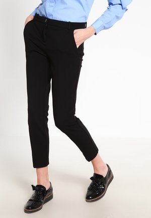 Person iført sort, tætsiddende bukser, lyseblå skjorte og skinnende sorte snørebåndssko, stående med hænderne i lommerne.