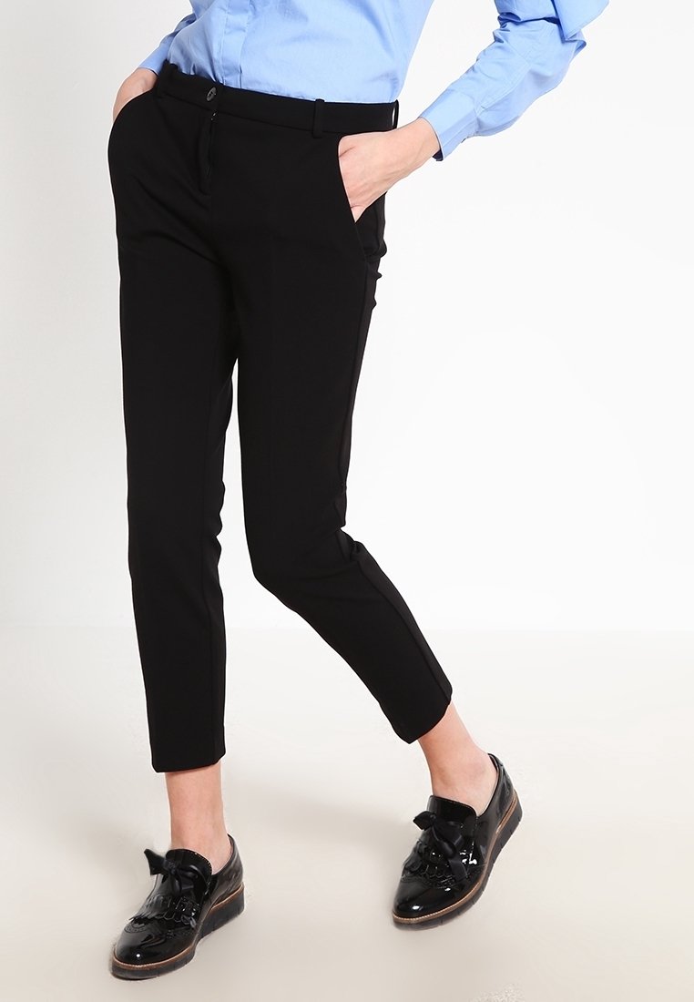 Personne portant un pantalon noir slim, une chemise bleu clair et des chaussures noires à lacets brillantes, debout les mains dans les poches.