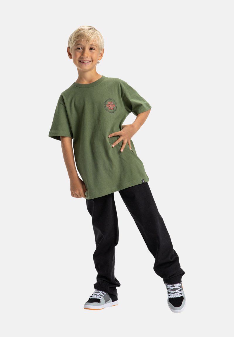 Zelené bavlněné oversized tričko s kulatým logem na hrudi, spárované s černými kalhotami a šedými teniskami s oranžovými akcenty.