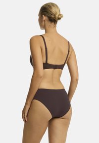 Modelka stoi plecami do kamery, ubrana w ciemno-brązowy biustonosz bikini oraz wysokie majtki, prezentując prosty i elegancki design.