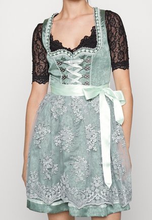 Femme portant une robe dirndl en satin vert avec un tablier en dentelle, des manches courtes en dentelle noire et un nœud en satin vert pâle noué à la taille.