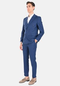 Completo blu a doppio petto con finitura testurizzata, pantaloni a sigaretta abbinati e scarpe in suede marrone chiaro. Camicia bianca sotto.