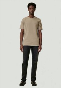 Ein Mann steht mit dem Gesicht nach vorne, trägt ein beiges T-Shirt mit kurzen Ärmeln, schwarze Jeans und schwarze Schuhe vor einem schlichten, hellen Hintergrund.