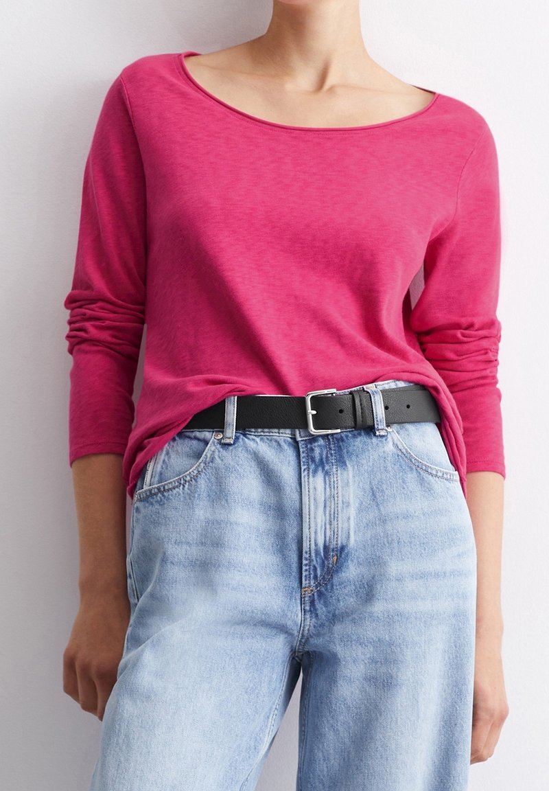 Kvinna som har på sig en långärmad klar rosa topp instoppad i ljusblå högmidjade jeans med ett svart bälte.