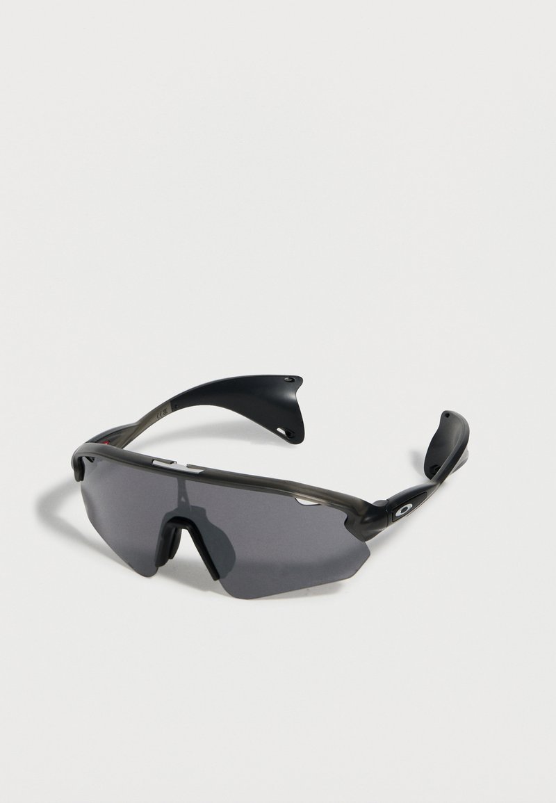 Μαύρα αθλητικά γυαλιά ηλίου με περιμετρικούς σκουρόχρωμους φακούς και κυρτές λαστιχένιες άκρες στα πλάγια, τοποθετημένα πάνω σε λευκή επιφάνεια.