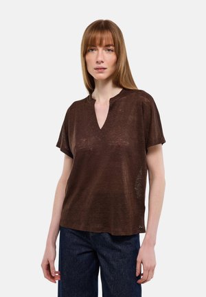 Femme aux cheveux roux lisses portant un haut ample à manches courtes marron et un jean bleu foncé, debout devant un fond clair uni.