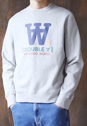 Osoba nosząca jasno szarą bluzę z niebieskim podwójnym logo W oraz tekstem "DOUBLE A BY WOOD WOOD", stoi z rękami w kieszeniach niebieskich dżinsów.