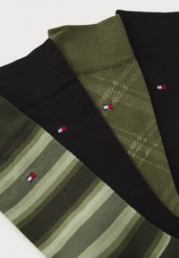 Tres pares de calcetines de lana: negros, verde oliva y a rayas verdes. Cada uno presenta un pequeño logotipo con detalles en rojo, blanco y azul.