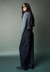 Sivý rebrovaný rolák v kombinácii s voľnými tmavými džínsami. Modelka má na sebe nadrozmerné zelené slnečné okuliare a čierne topánky.