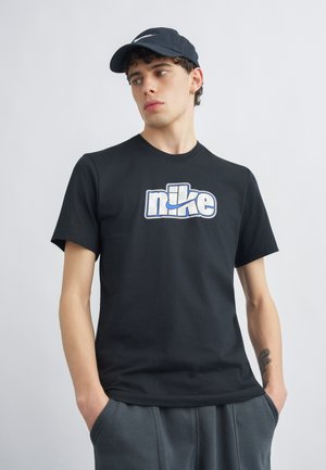 Fiatal férfi fekete Nike sapkában és fekete Nike pólóban fehér-kék logóval, zsebre tett kézzel, oldalra néz egy egyszerű háttér előtt.