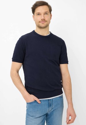 Homme portant un pull à manches courtes bleu marine avec une poche poitrine et un jean bleu clair, debout avec une main dans la poche sur fond blanc.