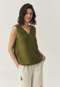 Top fără mâneci de culoare verde măsliniu cu un decolteu în V, realizat dintr-un material ușor, asociat cu pantaloni crem ce prezintă un detaliu de broderie cu albină.