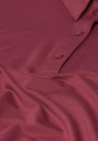 Burgunderfarbendes Poloshirt aus weichem Stoff mit Kragen und zwei passenden Knöpfen, sanft gefaltet ausgelegt.