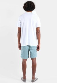 T-shirt blanc à manches courtes avec col rond, associé à un short bleu clair. Modèle portant des chaussures grises sans lacets, vu de dos.