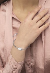 Bracciale d'argento con un charm ovale, design a catena linkata e piccole perle. Indossato su una mano, abbinato a un top in satin rosa chiaro.
