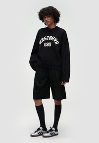 Czarny oversize sweter z białym napisem "WRSTBHV 030", czarne dopasowane szorty, czarne skarpety do kolan oraz srebrne trampki.
