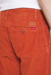 Pantalons en velours côtelé orange avec une poche arrière ornée d'une étiquette en cuir brun et d'une étiquette logo Levi's rouge cousue. Texture côtelée prononcée.