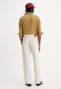 Lichtbeige wijde broek met een hoge taille en achterzakdetails, gecombineerd met een mosterdkleurig shirt en een rode pet, gezien vanuit de achterkant.