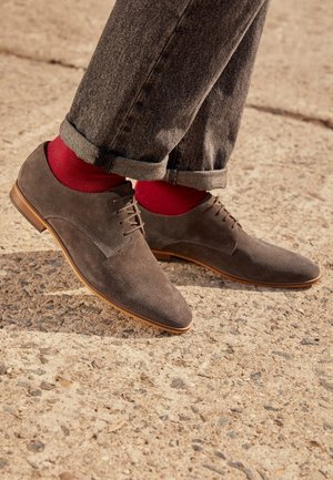 Chaussures habillées en daim marron avec un bout pointu et un design à lacets, associées à des chaussettes rouges et un jean gris foncé retroussé, sur une surface texturée.