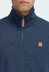 Muž oblečený v námorní modrom zipovacom saku s vysokým golierom a hnedým koženým ťahákom na zips a logom na hrudi.