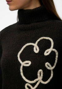 Czarny sweter dzianinowy z wysokim dekoltem, z wyraźnym kremowym wzorem kwiatowym wykonanym z grubego włóczki. Miękka tekstura z widocznym szyciem.