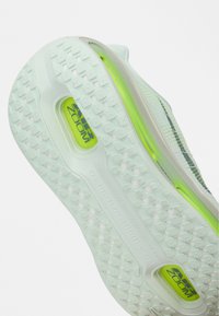 Talpa de pantof sport alb, cu o bază din cauciuc texturat, accente de culoare verde deschis pe mijloc și branding îndrăzneț "Air Zoom" în albastru închis.