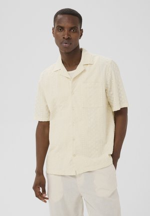 Mand med kort hår iført en cremefarvet, tekstureret kortærmet skjorte med knapper og brystlomme samt lyse bukser.