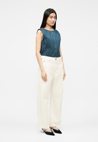 Γυναίκα που στέκεται φορώντας αμάνικο τοπ από denim, λευκό παντελόνι με φαρδιά μπατζάκια και μαύρες μυτερές γόβες σε απλό ανοιχτό φόντο.