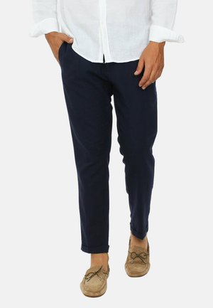 Parte inferiore del corpo di una persona che indossa pantaloni blu navy, mocassini beige e una camicia bianca a maniche lunghe con le maniche arrotolate.