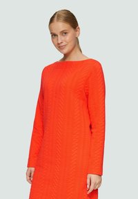 Orangenes langärmeliges Kleid mit Bootsausschnitt, das ein strukturiertes Zickzackmuster aufweist. Der Stoff wirkt dick und leicht dehnbar.