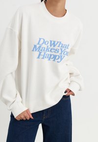 Sweatshirt blanc avec un col rond et une coupe oversize, arborant un texte bleu disant "Fais ce qui te rend heureux." Tissu doux, style décontracté.