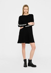 Zwarte jurk met lange mouwen en een uitlopende zoom, met witte merkaccenten op de mouwen. Gecombineerd met zwarte enkellaarsjes, tegen een neutrale achtergrond.