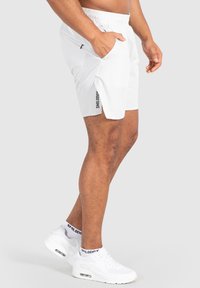 Pantalones cortos deportivos blancos con una cinturilla elástica, bolsillos laterales y un detalle mínimo de logo en negro en la pierna izquierda. Combinados con zapatillas blancas.