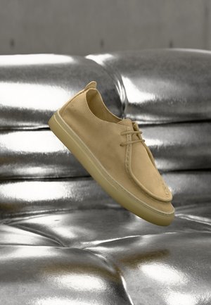 Zapato casual beige con cordones flotando sobre un fondo de cojín de sofá brillante de cuero plateado.