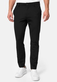 Zwarte slim-fit broek met een gladde textuur, voorzien van een knoopsluiting en zijzakken, gecombineerd met witte sneakers.