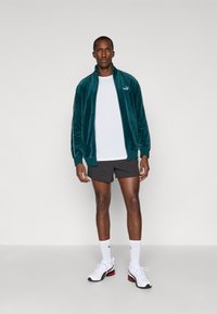 Téglazöld velúr sportdzseki, magas gallérral és cipzárral, fehér pólóval és szürke rövidnadrággal viselve; fehér sportcipővel és fehér zoknival párosítva.