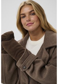 Jauna moteris dėvi rudą vilnonį paltą ir derančias pūkuotas pirštines, žvelgia į kamerą šiek tiek nusišypsojusi, priešais paprastą foną.