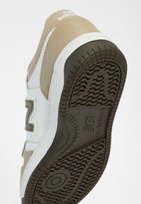 Baskets beige clair avec des accents blancs et une semelle extérieure texturée marron. Dotées d'un col rembourré et d'un logo "N" prononcé sur le côté.