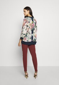 Blusa floral com base creme, apresentando padrões em rosa, roxo e verde, combinada com calças burgundy ajustadas e sandálias de salto nu.