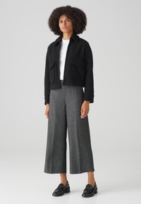 Zwarte cropped jacket met textuurstof en twee voorzakken, gecombineerd met grijze, wijde, geruite broeken en zwarte instapschoenen.