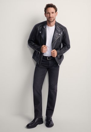Czarna skórzana kurtka motocyklowa z pikowanymi ramionami, biały t-shirt, ciemne dżinsy i czarne buty. Stoi na tle neutralnym.