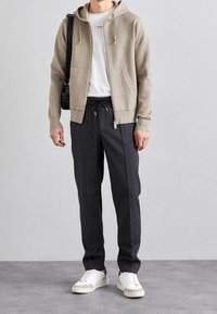 Beige Kapuzenpullover mit Reißverschluss über einem weißen Shirt, kombiniert mit dunkelgrauen Hosen und weißen Sneakern. Mit vorderen Taschen und Kordelzug in der Taille.