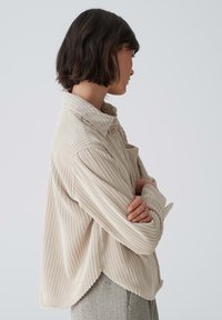 Beige corduroy shirt with a structured collar, ribbed texture, and a curved hem, worn with crossed arms, showcasing a relaxed fit.