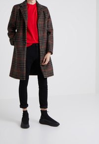 Brun rutig kappa med svarta och röda detaljer, bärs över en röd skjorta. Svarta åtsittande byxor och svarta slip-on skor kompletterar outfiten.