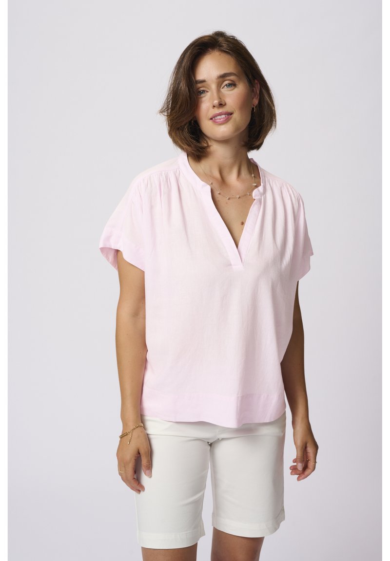 Blouse en lin rose clair avec des manches courtes, col en V sans boutons, et plis subtils aux épaules, assortie à un short blanc. Texture lisse.
