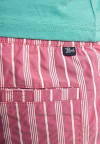 Maglietta turchese a maniche corte sopra pantaloni corti rosa con righe bianche verticali, dotata di una tasca posteriore e etichetta del marchio "Petrol".